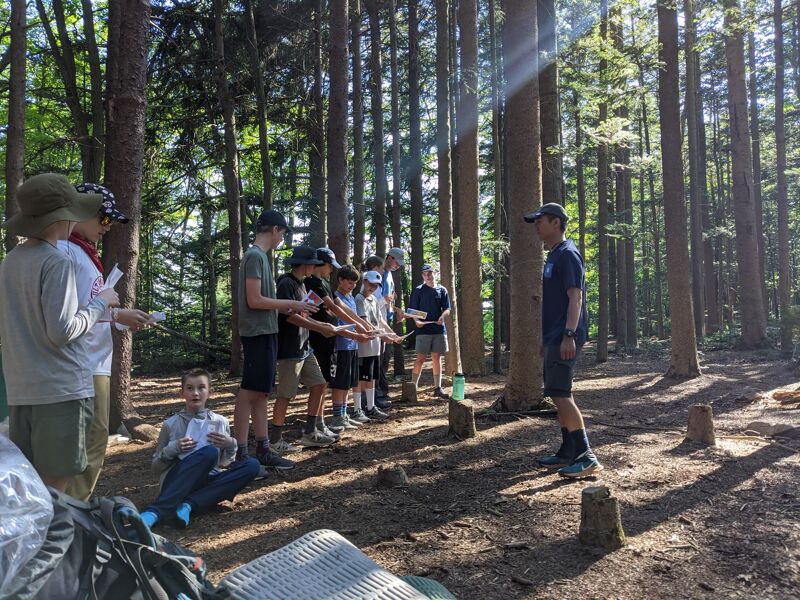 A group of people are standing in a forest, seemingly engaged in an activity or presentation. Some individuals are holding papers, while others are looking towards a person standing slightly apart from the group. The setting is a wooded area with tall trees and dappled sunlight filtering through the canopy. A person is sitting on the ground in the foreground. 